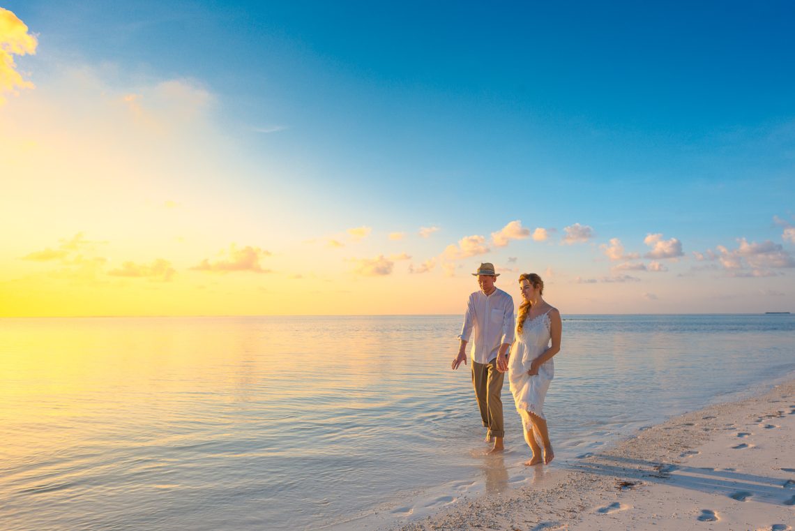 Couple walking on seashore wearing white tops during sunset