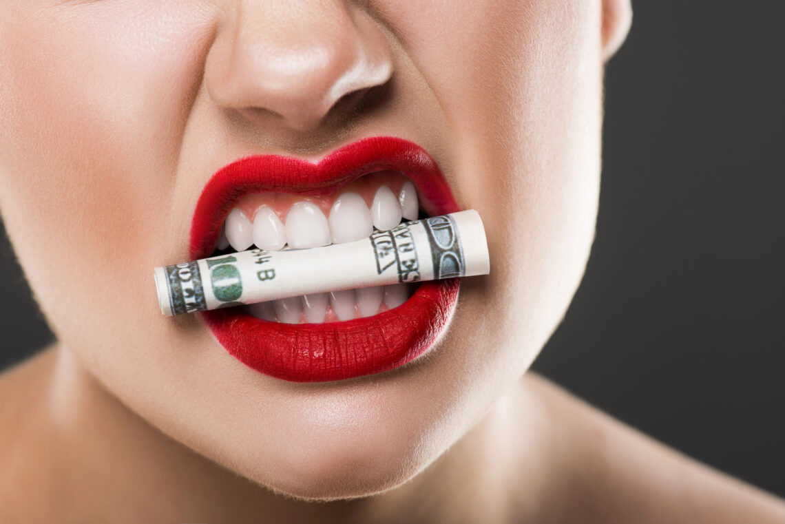 partial view of angry girl holding dollar banknote in teeth, isolated on grey