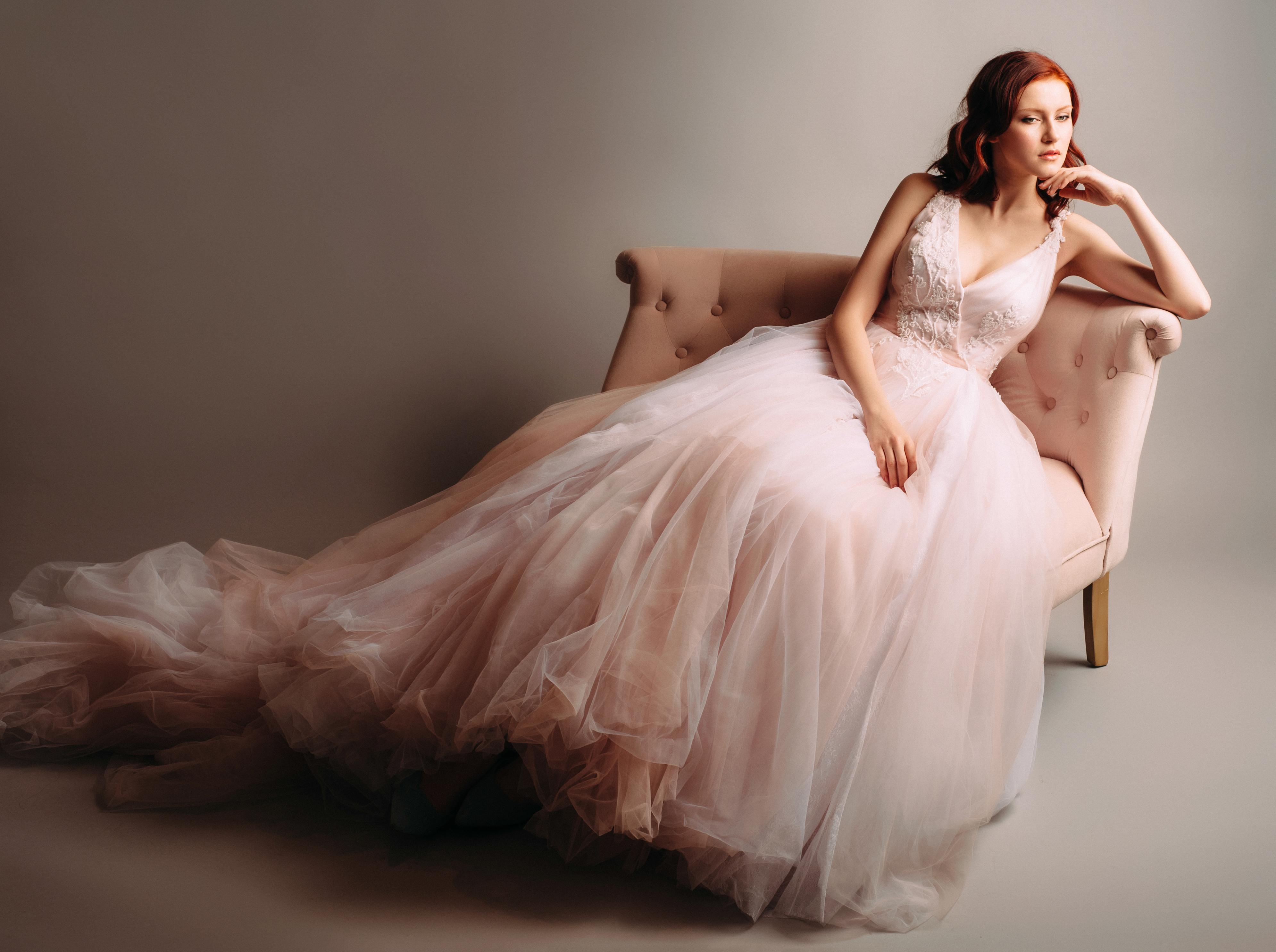 Redheaded bride in a pink wedding dress sitting in a vintage sofa. Studio portrait on neutral background.