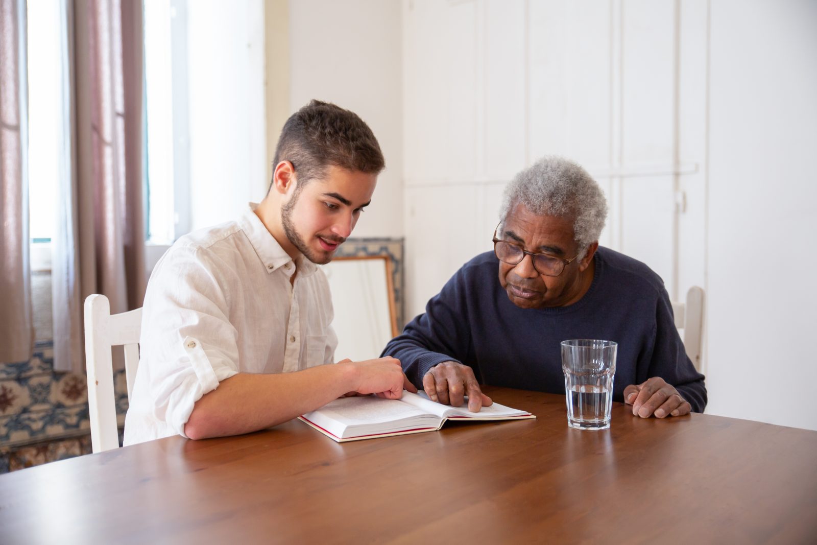 A man in white shirt talking to the man in blue sweater while reading a book