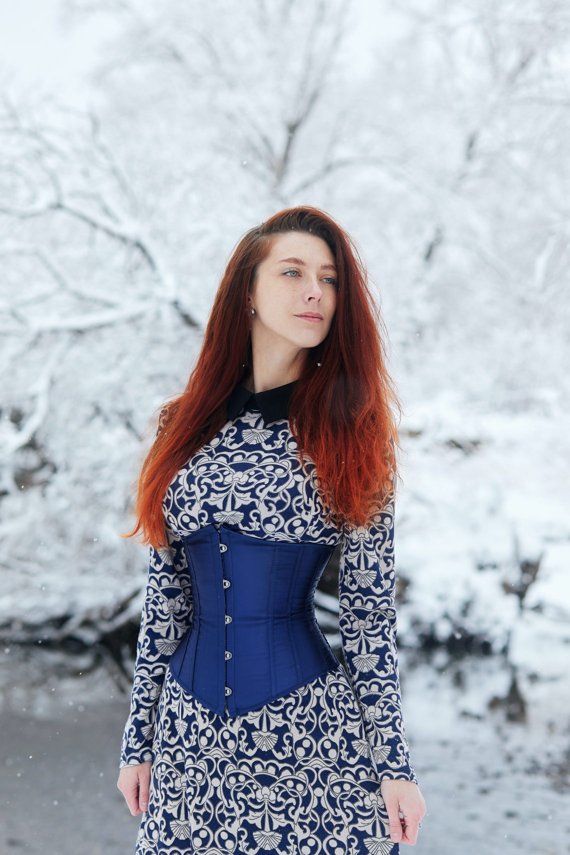 A spectacular winter portrait with a beautiful red-haired girl in a long dress and blue corset. Charming young woman posing in a snowy winter forest